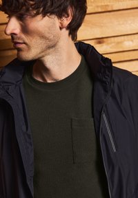 Černá bunda se zipem a límcem přes texturovanou tmavozelenou košili s náprsním kapsou. Dřevěné pozadí zvýrazňuje kompozici.