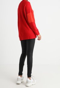 Pull rouge oversize à finition texturée, bande orange contrastante sur les manches, associé à des leggings noirs et des baskets blanches.