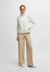 Gilet bianco trapuntato con colletto alto, chiusura a zip e tasche laterali, abbinato a pantaloni a gamba larga beige e sneakers bianche. Tessuto liscio.