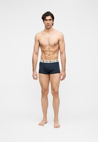 Νέος άνδρας με γυμνασμένο σώμα που φοράει μαύρα μπόξερ με λευκή μέση με πρόσωπα αρκούδων και το κείμενο "Moschino".