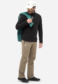 Schwarzer Fleece-Pullover, braune Hose mit Seitentaschen und schwarze Sneakers. Accessoire: türkisfarbener Mantel über der Schulter, weiße Mütze und Sonnenbrille.