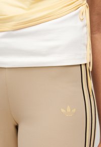 Μπεζ κολάν με μαύρες πλευρικές ρίγες και χρυσό λογότυπο Adidas. Η επάνω στρώση είναι ανοιχτό κίτρινο, με ένα λευκό φανελάκι να φαίνεται στο χείλος.