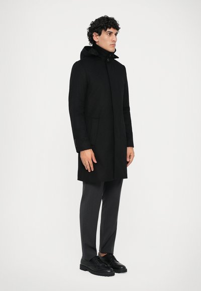 Abrigo negro con capucha, que cuenta con un tapeta frontal, bolsillos laterales y un ajuste entallado. Combinado con pantalones grises y zapatos negros.
