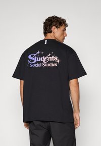 Svart bomulls t-skjorte med et stort flerfarget grafikk på baksiden som sier "Studenter Samfunnsfag" med stjerneakenter.