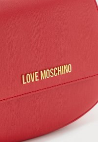 Piros műbőr keresztpántos táska sima felülettel. A táska elején feltűnően elhelyezett arany "LOVE MOSCHINO" logóval rendelkezik.