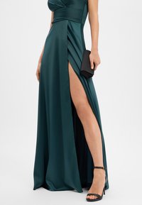 Femme portant une robe en satin vert foncé avec une fente haute sur la cuisse, tenant une pochette noire et portant des escarpins noirs à brides à la cheville.