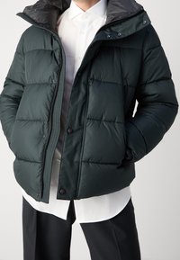 Uomo con giacca imbottita verde scuro sopra una camicia bianca a maniche lunghe e pantaloni neri, mani nelle tasche della giacca, su sfondo semplice.