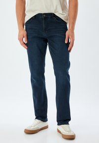 Jeans de mezclilla azul oscuro con un corte ajustado, diseño de cinco bolsillos, con un ligero desgaste. Combinados con zapatillas blancas, sobre un fondo simple.