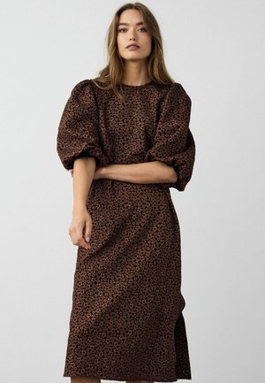 Frau in einem braunen, floral gemusterten Midikleid mit Puffärmeln, die vor einem schlichten hellgrauen Hintergrund steht.