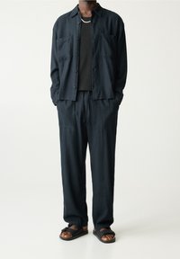 Donkerblauw shirt met twee borstzakken, gecombineerd met bijpassende losse broek. Zwarte t-shirt eronder en bruine sandalen met zwarte banden.