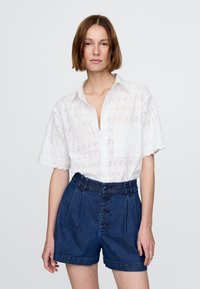 Balti marškiniai trumpomis rankovėmis su skylutėmis ir kišene, derinami su tamsios spalvos aukšto liemens džinsiniais šortais, kuriuose yra sagos priekyje ir klostės.