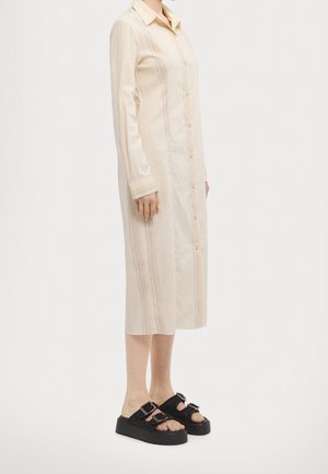Personne portant une robe midi beige à manches longues boutonnée et des sandales noires à plateforme ouvertes à l'avant avec des sangles à double boucle, debout de profil.