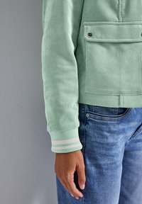 Mintgrüne Cord-Jacke mit vertikalen Rippungen, ausgestattet mit einer Fronttasche, weißen gestreiften Bündchen und kombiniert mit blauen Denim-Jeans.