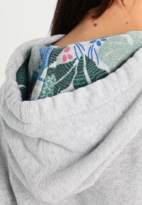 Sudadera con capucha gris que presenta un forro interior con un patrón floral, con detalles de costura a lo largo de los bordes y una textura suave.