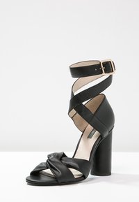 Musta nahkainen korkokenkä, jossa on solmittuja etuhihnoja ja ympärille kiedottava nilkkahihna, joka on kiinnitetty kultaisella soljella.