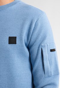 Lichtblauwe trui met een ronde hals, geribbelde manchetten en een klein zwart logo op de borst en mouw. Zachte, gestructureerde stof.