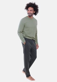 Olivegrünes Langarmshirt kombiniert mit schwarzen Loungehosen mit Muster, ausgestattet mit einem elastischen Bund und einem lockeren Schnitt. Barfuß vor einfachem Hintergrund.