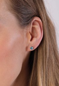 Ronde studoorbellen met een lichtblauwe edelsteen in een metalen zetting, gedragen op een lichte oor met steil bruin haar.