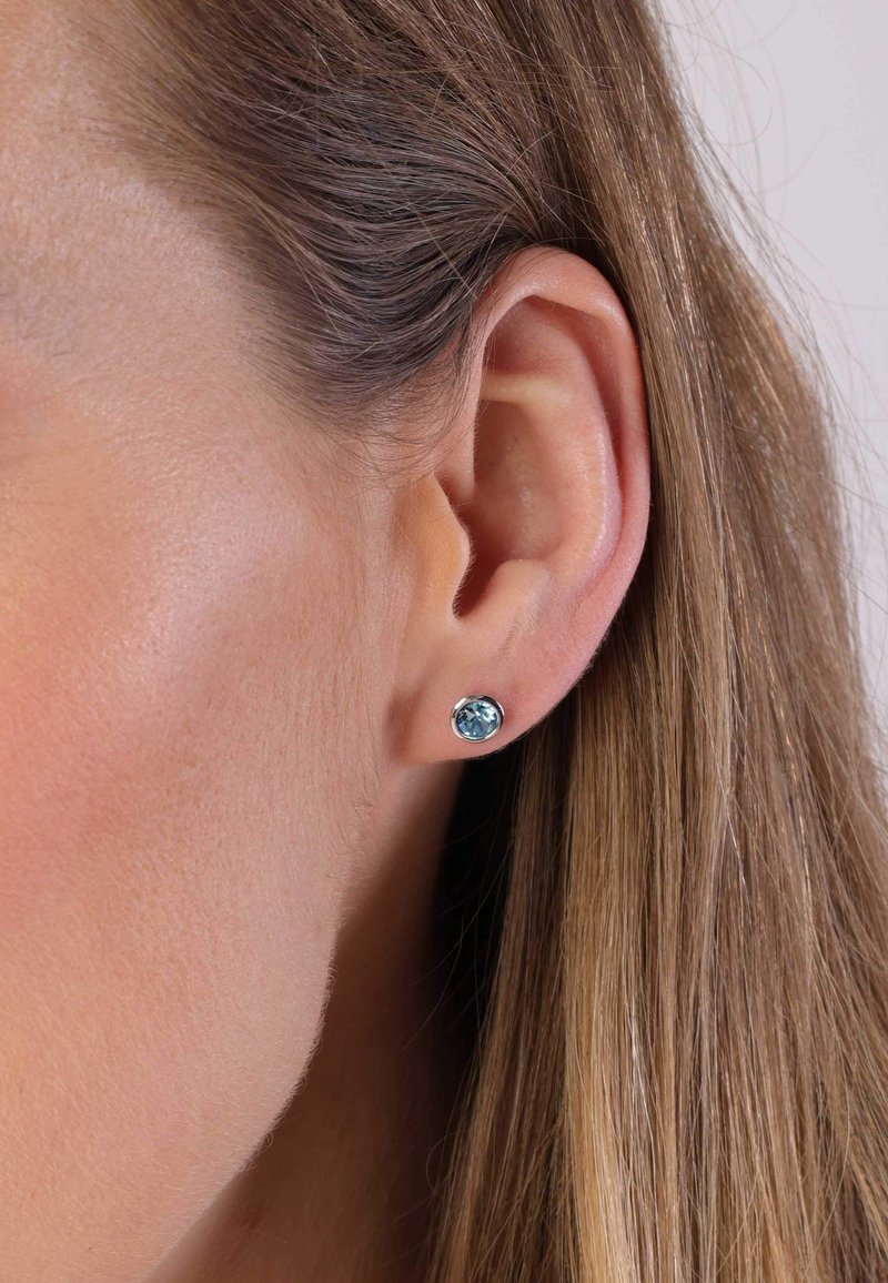 Ronde studoorbellen met een lichtblauwe edelsteen in een metalen zetting, gedragen op een lichte oor met steil bruin haar.