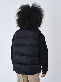 Chaleco de plumas negro con una textura suave y acolchada, que presenta un cuello alto y una capucha. El chaleco se combina con una camisa negra de manga larga.
