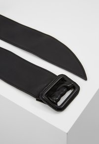 Svart skinnbelte med en glatt overflate, med en rektangulær metallspenne og sydd kanting. Formen er avlang og avsmalnende i den ene enden.