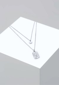 Deux colliers en argent posés sur un cube blanc ; l'un avec un pendentif rond lisse, l'autre avec un pendentif rectangulaire texturé.