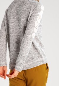 Grå stickad tröja med texturerad yta, med en blommig spetsdetalj längs vänster ärm. Kombinerad med bruna byxor.