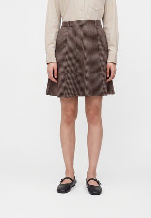 Person, die ein beiges Langarmshirt, einen braunen Hahnentrittmuster A-Linien-Rock und schwarze Mary Jane Ballerinas trägt, steht vor einem schlichten Hintergrund.