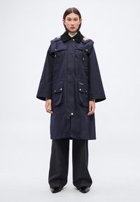 Μπλε σκούρο αδιάβροχο παλτό με ψηλό γιακά, τέσσερις μπροστινές τσέπες, αντιθετικές λεπτομέρειες στους ώμους και χρυσούς μεταλλικούς εξαρτήματα. Φαρδιά μανίκια και ίσια γραμμή.