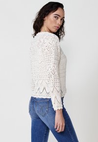 Suéter de crochet crema con patrones de diamante y olas, mangas largas y bordes con forma de concha, combinado con jeans ajustados azules.