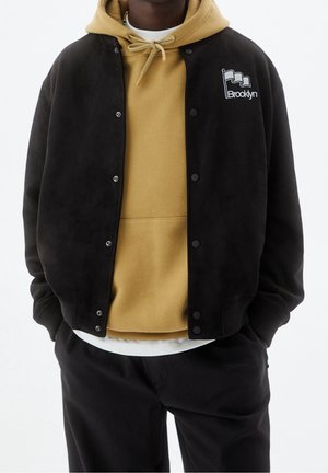 Persoon draagt een zwarte jas met drukknopen en een Brooklyn-logo over een mosterdgele hoodie en zwarte broek, handen in de zakken.