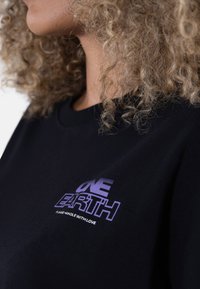 Svart bomull t-skjorte med et lilla grafisk tekst "ONE EARTH" og frasen "PLEASE HANDLE WITH LOVE" på venstre brystområde.