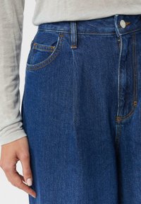 Niebieskie dżinsy z denimu mają wysoki stan, pomarańczowe przeszycia i przednią kieszeń. Faktura jest gładka z delikatnym połyskiem.