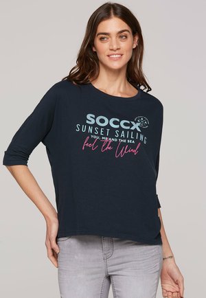 Navy blue, loose-fitting t-shirt with three-quarter sleeves. Front features a graphic print in light blue and pink with text and logo accents.