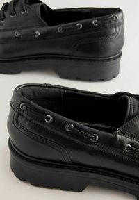 Scarpe in pelle nera con design allacciato, finitura testurizzata, suole in gomma e occhielli decorativi lungo i lati. Tacco svasato per maggiore stabilità.