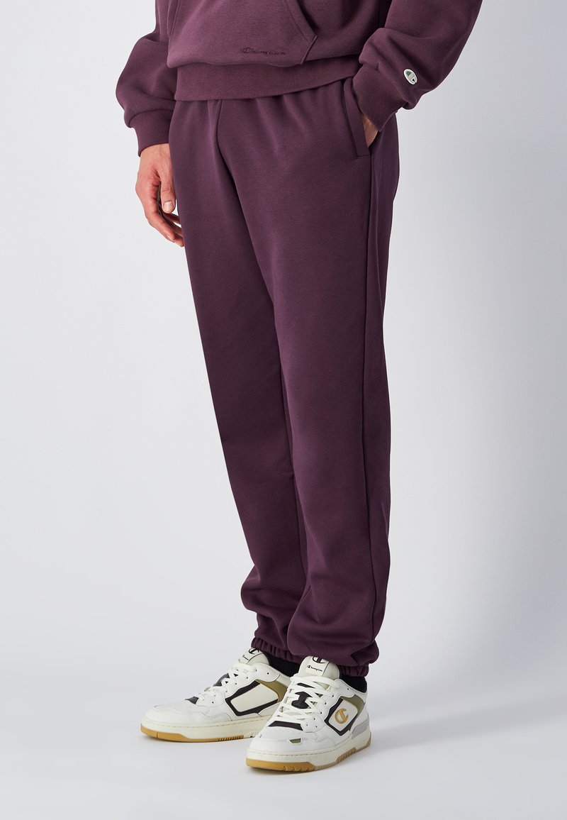 Verbrande paarse joggingbroek met een relaxte pasvorm, elastische tailleband en zijzakken, gecombineerd met wit-gouden sportschoenen.