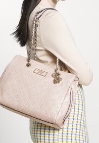 Ljusrosa handväska med präglat logomönster, guldfärgade detaljer och dubbla kedjehandtag. Dragkedjedetaljer i sidan ger struktur.