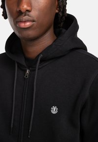 Schwarze Zip-Hoodie aus weichem Stoff mit einem vorderen Reißverschluss, Kordelzügen und einem weißen Logo-Emblem auf der Brust.