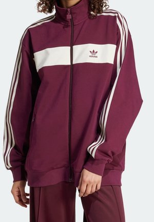 Personne portant une veste de survêtement Adidas bordeaux avec des bandes blanches sur les manches et une bande blanche horizontale sur la poitrine.