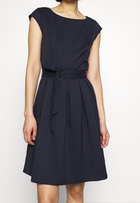 Žena v bezrukávových námorně modrých šatech s texturou, s lodičkovým výstřihem a uvázaným látkovým páskem v pase, stojící na jednobarevném pozadí.