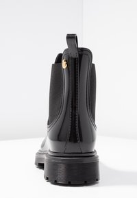 Botín negro con un acabado brillante de charol, paneles laterales elásticos, suela de goma, costuras en contraste y un pequeño acento dorado en el tacón.