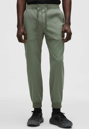 Mand iført olivengrønne joggingbukser med snøre i taljen og sorte sportssko, stående mod en ensfarvet baggrund.