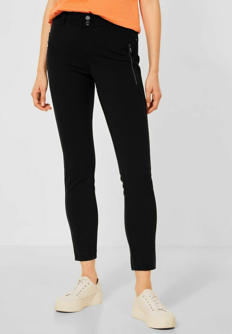 Černé slim kalhoty s dvěma předními zipovými kapsami, hladkou texturou a úzkým střihem. Kombinované se světlými teniskami.