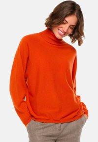 Pull léger orange en col roulé, fabriqué dans un tissu doux, avec une coupe décontractée et des manches longues, associé à un pantalon à motifs.