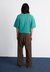 Tricou din bumbac turcoaz, cu mâneci scurte și o croială lejeră, asortat cu pantaloni largi maro. Vedere din spate; design minimalist, culori solide.