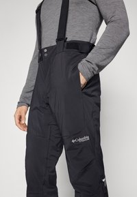 Pantalons de neige imperméables noirs avec bretelles réglables, boutons à l'avant, poches et logo Columbia Titanium sur la jambe inférieure.