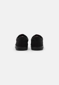 Zapatillas negras con textura suave, parte trasera redondeada y suela de goma en contraste. Presentan el logotipo de la marca en el talón y acentos rojos en la lengüeta.