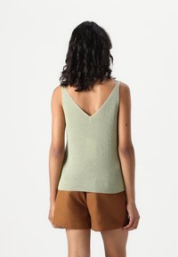 Grønn, ermeløs strikket topp med V-hals og subtil glans, sammen med brune shorts. Glatt tekstur, slank passform og lengde under hoften.