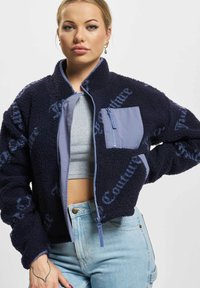 Mlada ženska nosi temno modro flis jakno z zadrgo na prsnem žepu, oblečeno čez svetlo sivo kratko majico in svetlo modre kavbojke, stoji s svojo roko na boku.