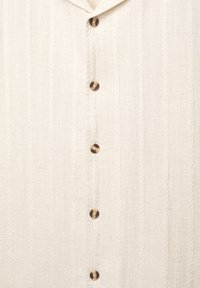 Cream button-up shirt made of textured fabric, featuring vertical stripes and dark buttons along the front.